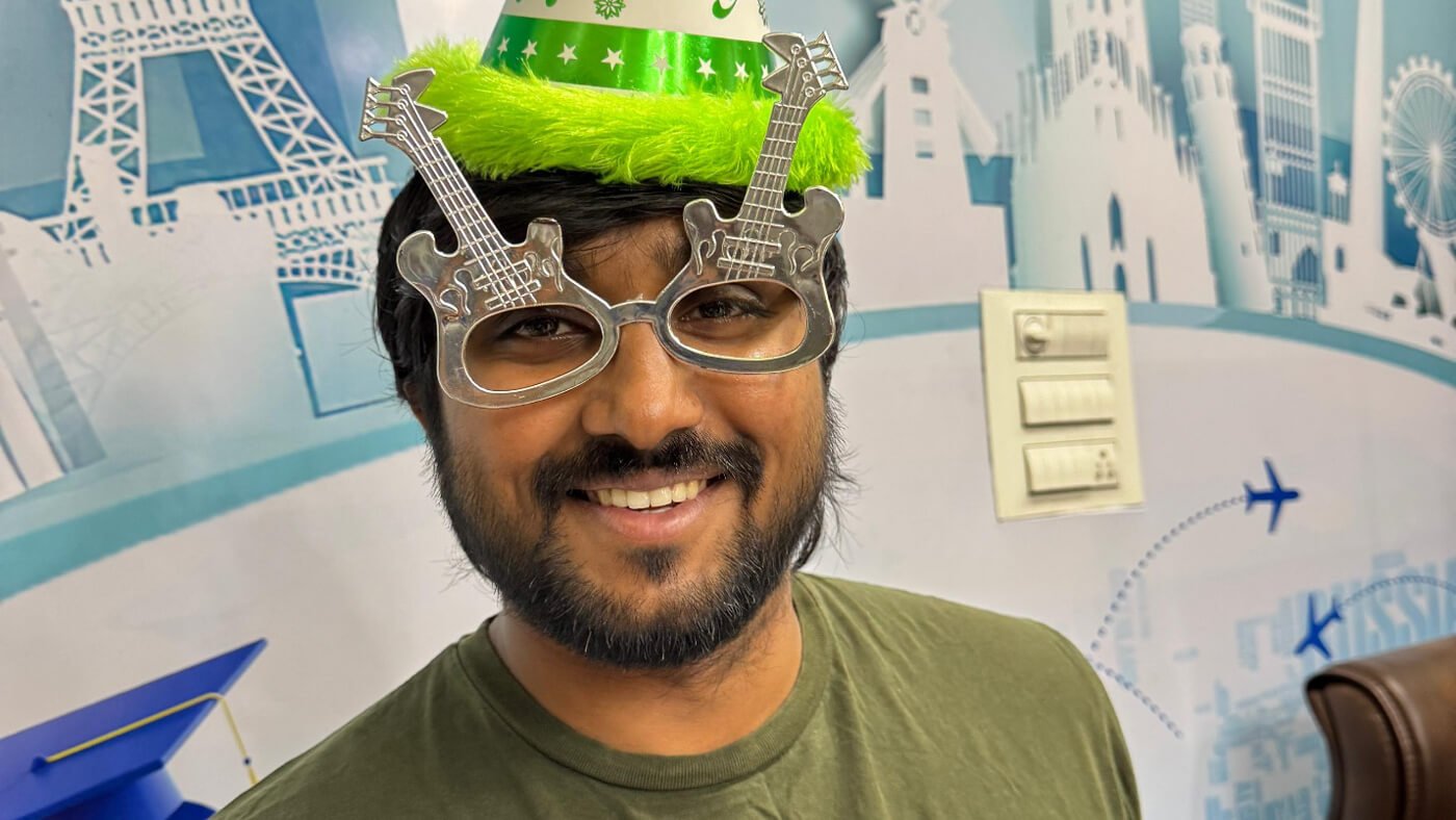 A smiling person wears novelty guitar-shaped glasses and a festive green party hat with a fluffy brim, standing in front of a world landmark-themed backdrop.