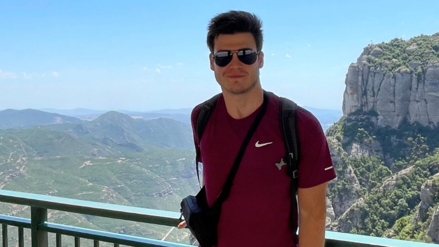 A man wearing sunglasses and a maroon shirt stands at a scenic overlook with vast mountain ranges behind him under a bright blue sky.
