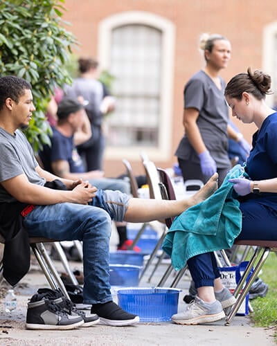 A medical student provides foot care to a seated patient during an outdoor community clinic, while other clinicians and patients are visible in the background.