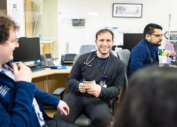 Medical residents gather in a clinical workroom, collaborating and reviewing information at computer workstations.