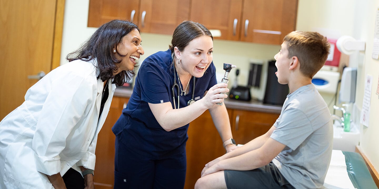 A medical student uses an otoscope to examine a child’s throat in a medical exam room while another clinician observes.