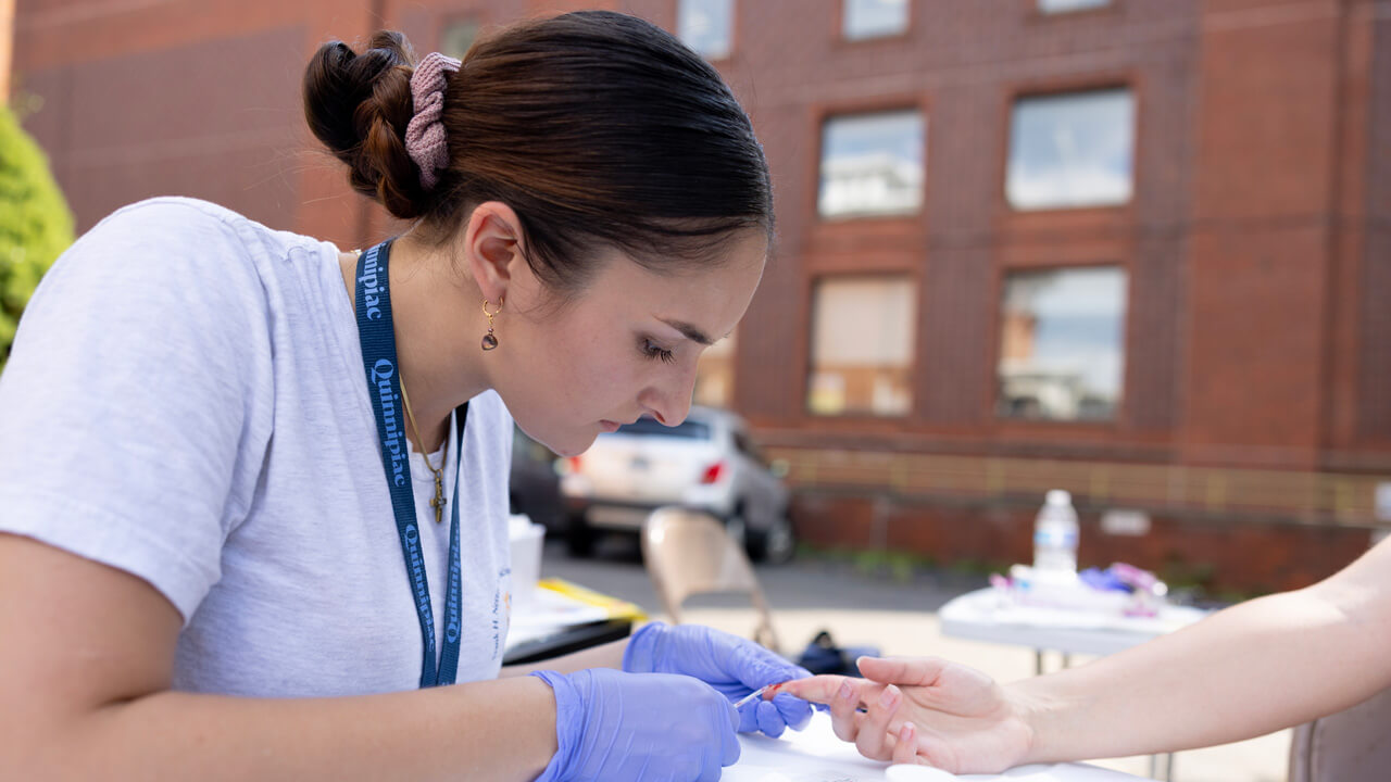 A female medical student collects a blood sample from a patient's finger at an outdoor clinic table.