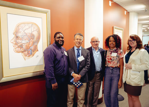5 people smile in front of Frank Netter's illustration of a human skull and nervous system