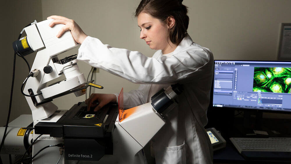 A medical student wearing a lab coat uses a large machine to process lab samples in the research laboratory