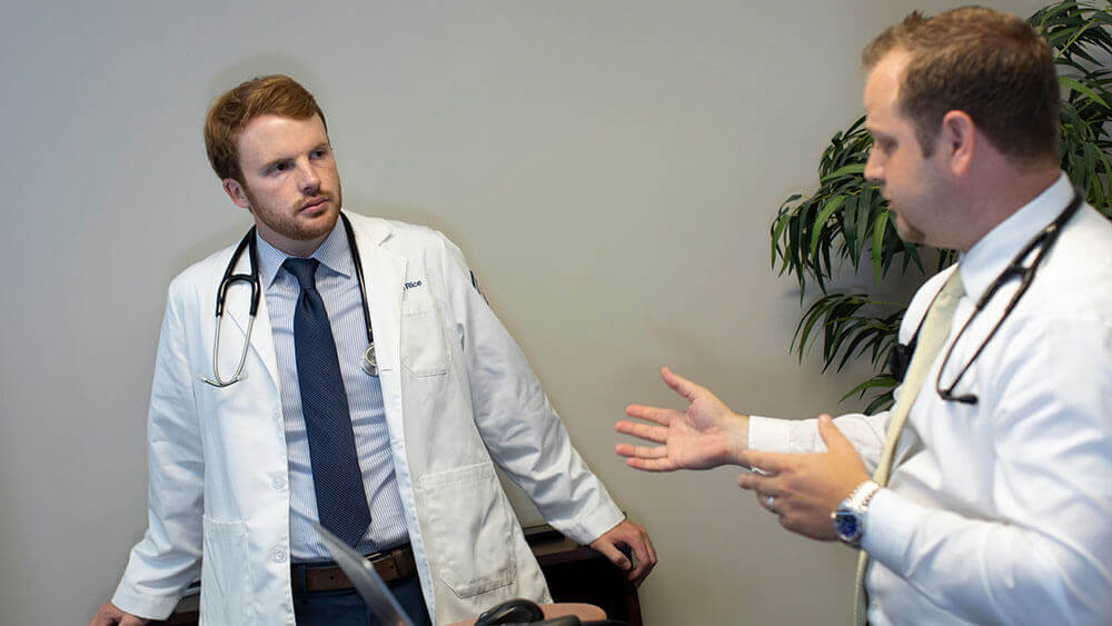 A medical student wearing a stethoscope speaks to his mentor doctor at his medical practice