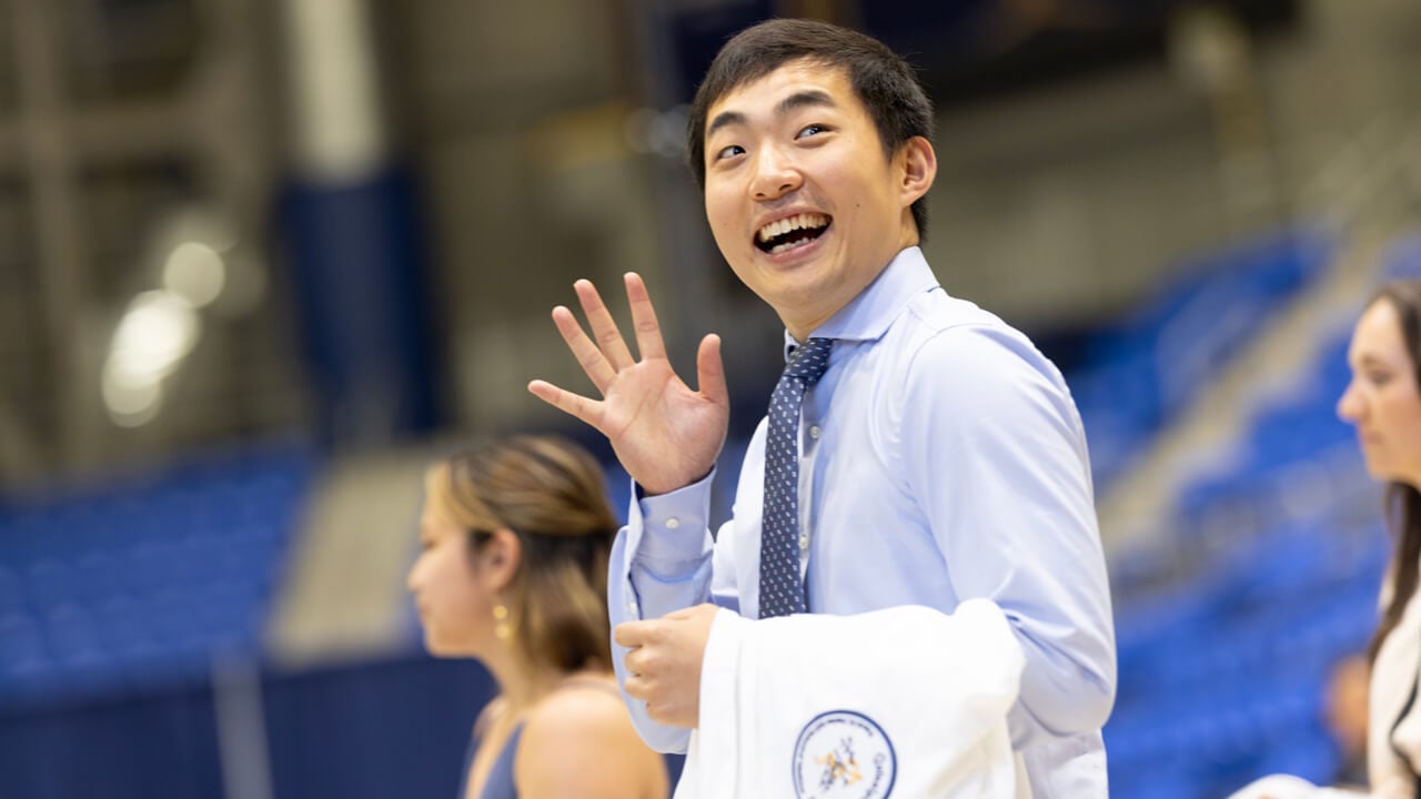 A medical student smiles broadly and waves from the stage